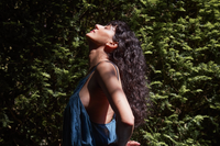 A person with long, curly dark hair stands outdoors in front of dense green foliage, tilting their head back and closing their eyes, bathed in dappled sunlight and wearing a sleeveless blue top.