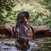 A person with wet, dark hair relaxes in an outdoor wooden tub, resting their arms on the edge and facing a lush, green, natural background.