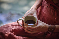 A person in a reddish-brown dress holds a ceramic mug filled with black coffee, sitting outdoors with soft natural light.