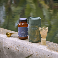 A jar labeled The Hydration, a cylindrical container labeled Matcha Collagen, a gold measuring spoon, and a bamboo whisk sit on a stone ledge beside a pond with greenery reflected in the water.
