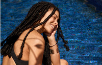 A woman with long, black locs sits by a pool, smiling with her eyes closed and her hand resting on her cheek. She wears a black swimsuit and bracelets, with sunlight casting shadows on her skin.