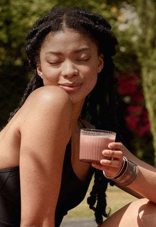 A woman in a black swimsuit sits by a pool, eyes closed and smiling softly, holding a glass of a pink drink. The background features lush greenery and colorful blooming shrubs.