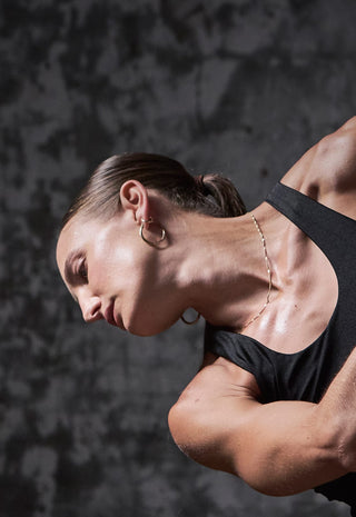 A woman wearing a black sports bra and jewelry poses with her arms extended and head tilted, against a dark, textured background.