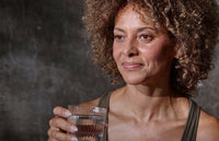 A woman with curly hair holds a glass of water and smiles softly. She is wearing a sleeveless top and is positioned against a dark, textured background.