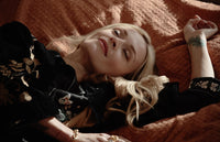 A woman with long blonde hair lies on an orange textured blanket, eyes closed and relaxed, wearing a black floral jacket. Her right arm is raised above her head, and soft light highlights her peaceful expression.