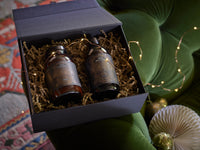 A gift box with two dark glass jars filled with shredded paper sits on a green velvet cushion, surrounded by festive string lights and holiday decor.