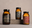 Three amber jars with decorative labels, including black, red, and orange designs, are displayed on a light brown surface and stone block against a neutral background. The jars appear to be food or spice products.