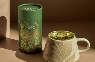 A green canister labeled Ancient + Brave Matcha + Collagen stands next to a ceramic cup filled with frothy matcha. Both are on a beige surface with artistic lighting and shadows.
