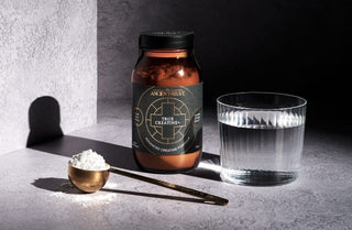 A jar labeled True Creatine+ stands next to a glass of water and a gold scoop filled with white powder on a gray surface with dramatic lighting and shadows.