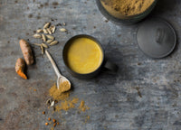 A black mug filled with golden turmeric latte sits on a gray surface. Nearby are cardamom pods, turmeric root, a spoon of ground spice, scattered powder, and an open jar of spice powder.