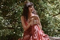 A woman in a flowing pink dress sits outdoors on a wooden surface, holding a mug with both hands, surrounded by lush green foliage and dappled sunlight.