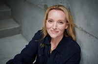 A woman with blonde hair wearing a dark jacket sits and smiles gently, leaning against a gray concrete wall with stairs in the background.