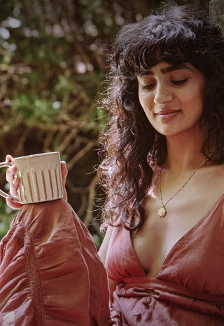 A woman wearing a rust-colored dress stands outdoors, holding a striped mug and smiling softly. Sunlight filters through greenery in the background.
