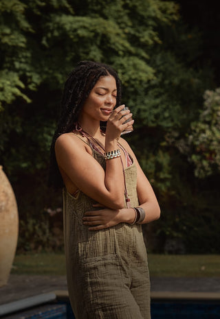 A woman with long braided hair stands outdoors, eyes closed, peacefully holding and smelling a glass. Lush green foliage and an urn are visible in the background.