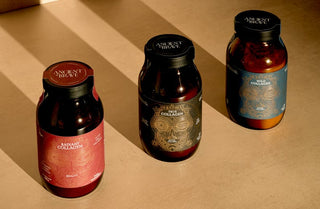 Three jars of Ancient + Brave collagen supplements in red, blue, and brown labels are placed on a beige surface, illuminated by warm sunlight and casting diagonal shadows.