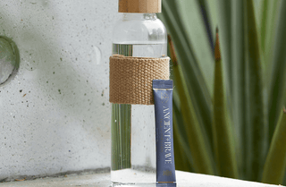 A clear glass water bottle with a tan woven sleeve and cap stands on a stone surface next to a blue Ancient + Brave drink sachet. Green, spiky plant leaves are blurred in the background.