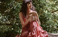A woman with wavy dark hair sits outdoors in front of leafy green plants, wearing a flowing rose-colored dress and holding a mug, gazing thoughtfully to the side.