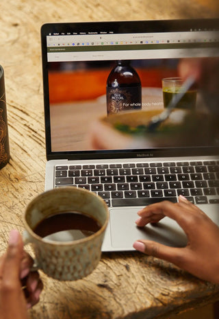 A person holding a mug of coffee uses a laptop on a rustic wooden table. Next to the laptop is a container of Ancient Coffee Company coffee, and the laptop screen displays a webpage featuring the same product.
