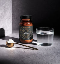 A jar labeled True Creatine, a glass of water, a gold measuring spoon, and a heap of white creatine powder sit on a textured grey surface, illuminated by natural light.