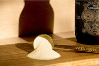 A gold scoop labeled ANCIENT + BRAVE holds and spills a mound of white collagen powder onto a wooden surface, with a dark container labeled True Collagen nearby.