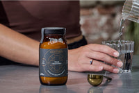 A person pours water into a glass beside a brown jar labeled True Creatine+ and a gold measuring scoop on a silver countertop. The person wears a brown top and a ring on their finger.