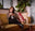 A woman sits on a mustard-yellow sofa, smiling and holding a mug. She is surrounded by patterned cushions and plants, with a coffee table and artwork visible in the cozy, warmly lit living room.