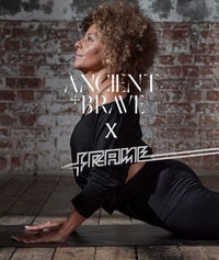 A woman with curly hair practices yoga indoors, performing an upward dog pose on a mat. Behind her is a rustic brick wall. Text on the image reads ANCIENT + BRAVE X FRAME.
