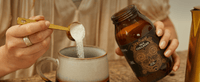 A person pours a scoop of white powder from a jar labeled Wild Collagen into a ceramic mug on a wooden table.