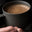 A hand in a dark sweater holds a matte black mug filled with a creamy, frothy hot beverage, possibly coffee or hot chocolate, against a dark, blurred background.