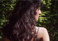 A woman with long, wavy dark hair stands outdoors, turned slightly to the side, with green foliage in the background and sunlight highlighting her hair and shoulder.