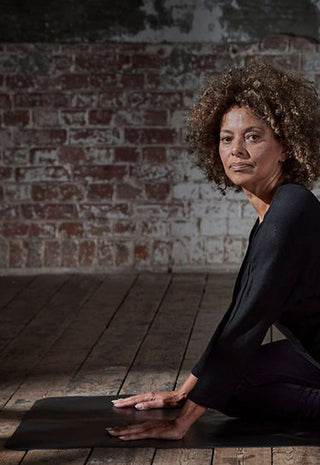 A woman with curly hair wearing black athletic clothing practices yoga on a mat in a dimly lit room with wooden floors and an exposed brick wall, performing a seated spinal twist pose.