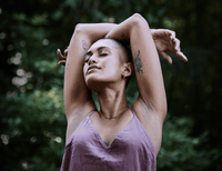 A person with short hair and visible tattoos stretches their arms above their head, eyes closed, wearing a sleeveless mauve top, standing outdoors with a blurred green background.