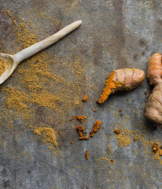 A wooden spoon with ground turmeric, fresh turmeric root, and cardamom pods are scattered on a rough metal surface.