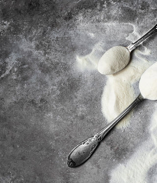 Three vintage silver spoons filled with white powder, possibly flour or powdered sugar, are scattered on a textured gray surface, with some powder spilled around them.