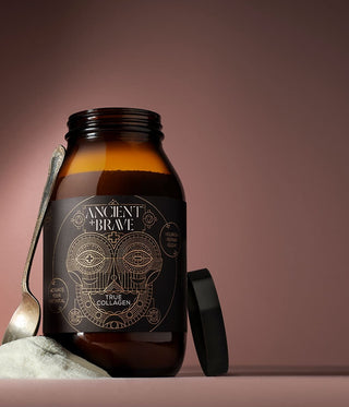 A brown glass jar labeled Ancient + Brave True Collagen stands on a surface with a spoon and a small mound of powder nearby, against a gradient brown background. The jars black lid rests beside it.