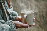 A person wearing a green jacket holds a steaming ceramic mug with both hands outdoors, with a blurred natural background.