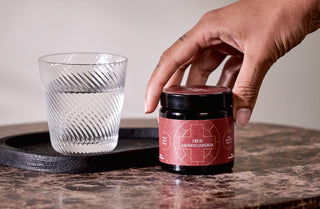 A hand reaches for a jar labeled True Ashwagandha on a marble table, next to a textured glass of water on a black tray.