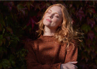 A woman with long red hair stands against a background of dark leafy vines, eyes closed and face tilted up, enjoying the sunlight. She wears a brown sweater and looks peaceful and relaxed.