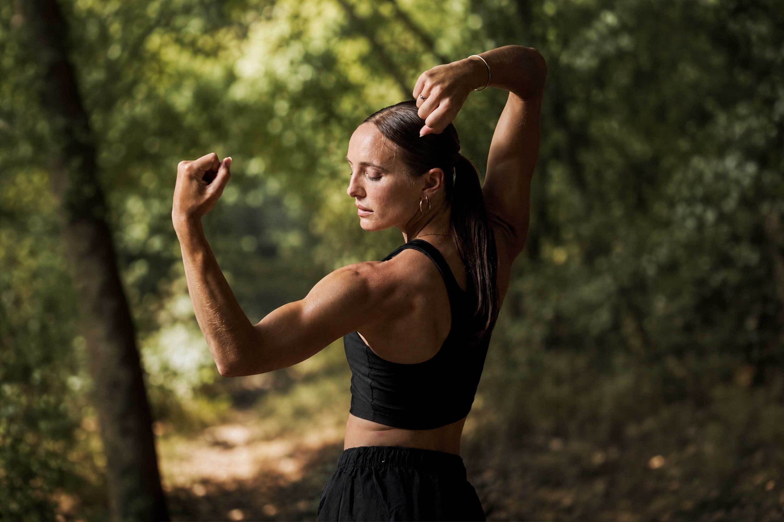 woman with brown hair flexes arms in the forest with light and shadow casting over her arms and face