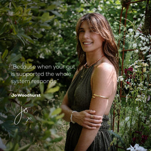 A woman in a sleeveless dress stands among flowers, smiling with arms crossed. Text reads: With True Biome by Ancient + Brave US, strong gut support means your whole system responds. — Jo Woodhurst.