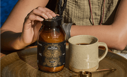 A person holds a jar labeled True Collagen on a brass tray beside a beige mug filled with a dark drink and a gold measuring spoon, with sunlight casting shadows.