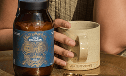 A person holding a large beige mug sits behind a jar labeled Ancient + Brave Wild Collagen on a gold tray, with a gold measuring spoon nearby. The person is wearing a textured, earth-toned garment.