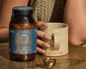 A person holding a large beige mug sits behind a jar labeled Ancient + Brave Wild Collagen on a gold tray, with a gold measuring spoon nearby. The person is wearing a textured, earth-toned garment.