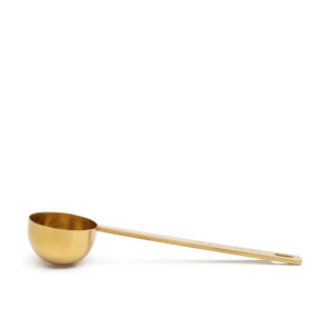 The Ancient + Brave Ritual Scoop, a golden measuring spoon with a long handle, is displayed on a flat brown surface against a light background.