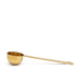 The Ancient + Brave Ritual Scoop, a golden measuring spoon with a long handle, is displayed on a flat brown surface against a light background.