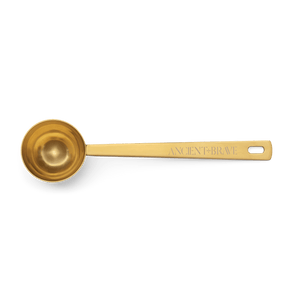 The Ritual Scoop by Ancient + Brave is a golden stainless steel scoop with ANCIENT + BRAVE engraved on the handle—a stylish wellness gift, beautifully displayed on a plain background.