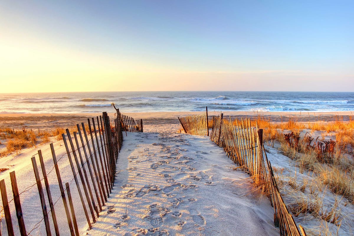 A sandy path with rustic wooden fences leading from the dunes to the ocean at sunset, with waves rolling in under a clear blue sky.