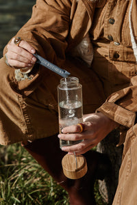 A person in a brown outfit sits outdoors, holding a glass water bottle and pouring powder from a packet labeled “ANCIENT B R W” into it. The bottle’s wooden lid rests in their other hand. Grass is visible in the foreground.
