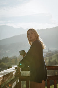A woman with long blonde hair stands on a wooden balcony, holding a mug and gazing towards the camera. She is wearing a dark oversized sweater, with misty mountains and fields in the background on a bright sunny day.