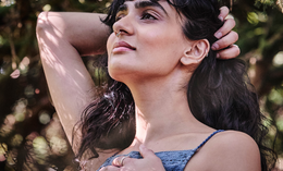 A person with long dark hair and a nose ring stands outdoors, one hand behind their head and the other on their chest, gazing upward with sunlight and tree shadows on their face.
